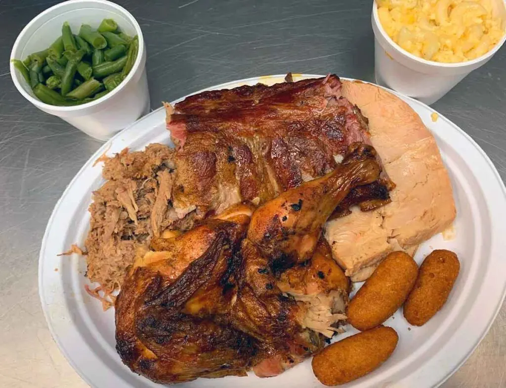 Plate filled with BBQ chicken, ribs, turkey, and pork. Two cups on side. One with green beans, the other with Mac n cheese.