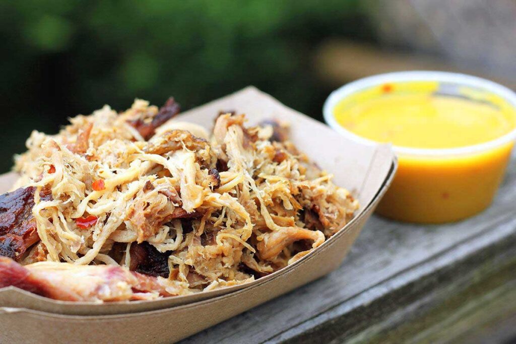 Closeup of paper tray of pulled pork beside plastic container of mustard sauce in outdoor setting.