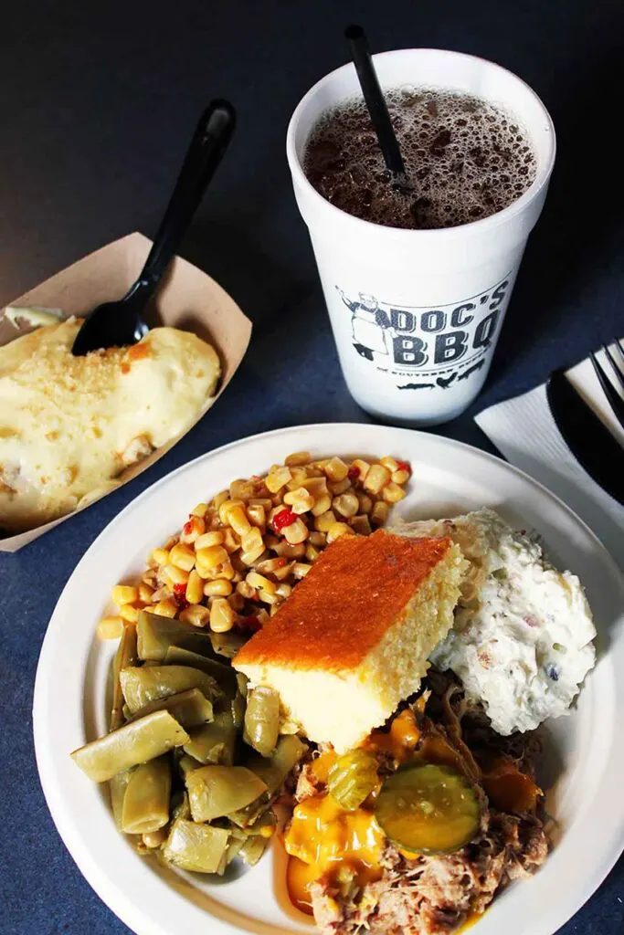 Plate of BBQ, beans, corn, and other sides with drink on table.