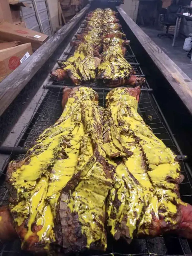 Looking long ways down a long pit filled with whole hogs covered in mustard BBQ sauce