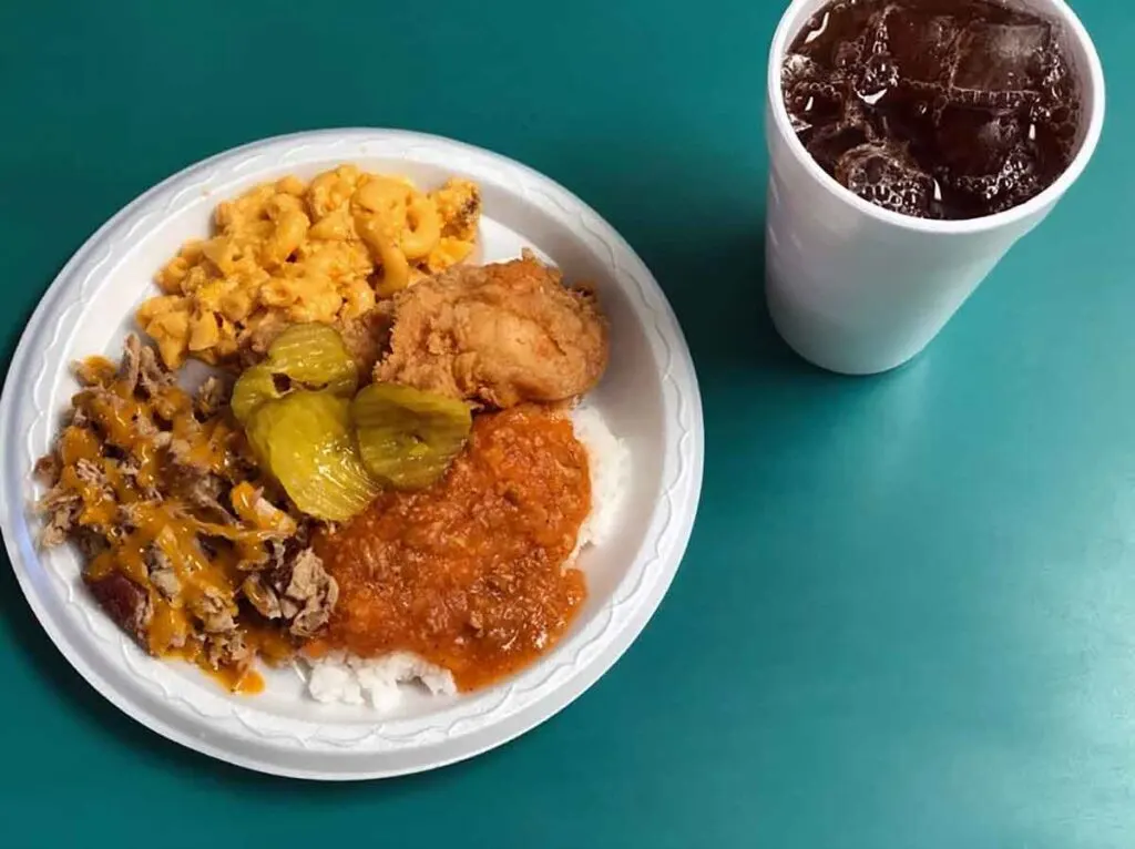 Plate of BBQ, hash, fried chicken, Mac and cheese, and pickles on green tabletop with cup full of iced tea.