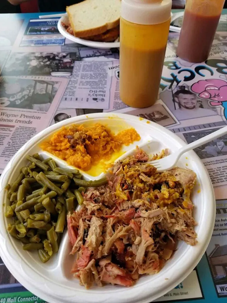 Plate of pulled pork, green beans, and sweet potato casserole with bottles of sauces and bread in background.