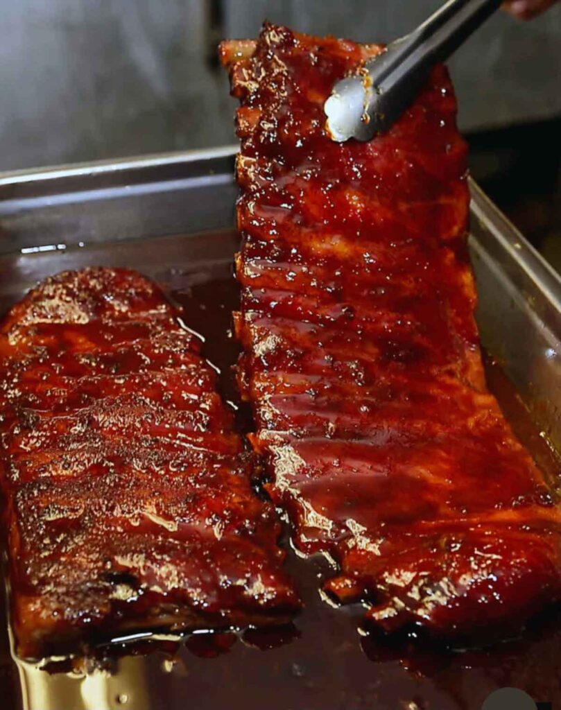 Two racks of ribs in sauce, one being held up on one end by tongs