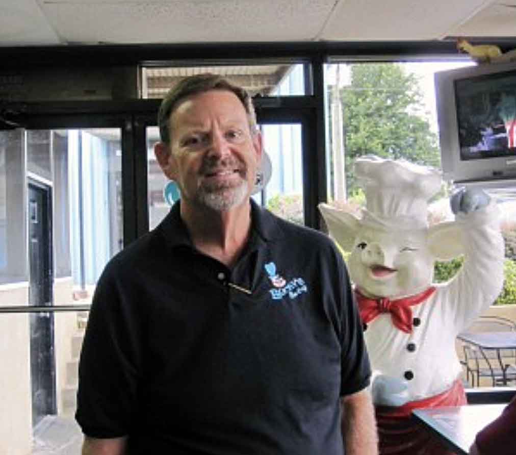 Wayne Preston, owner of Bucky's BBQ, with smiling white pig statue waving in the background.