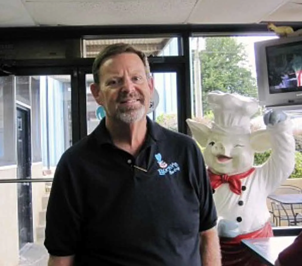 Wayne Preston, owner of Bucky's BBQ, with smiling white pig statue waving in the background.