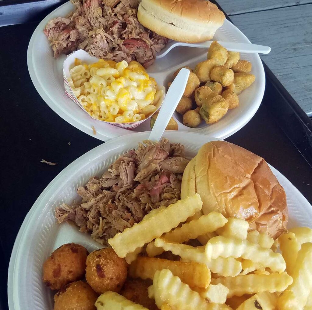 Two plates of BBQ sandwiches with sides, including fries, Mac and cheese, fried okra and hushpuppies.