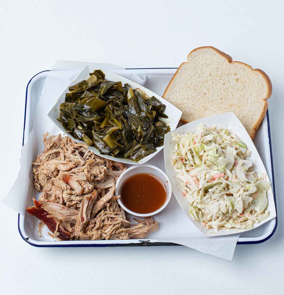 Whole hog bbq plate with collards, slaw, sauce and bread on tray.