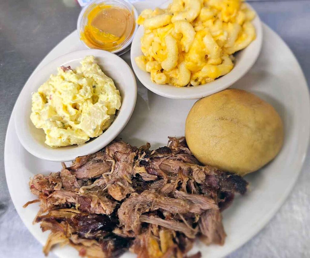 Plate of pulled pork, potato salad, Mac and cheese, with a roll.