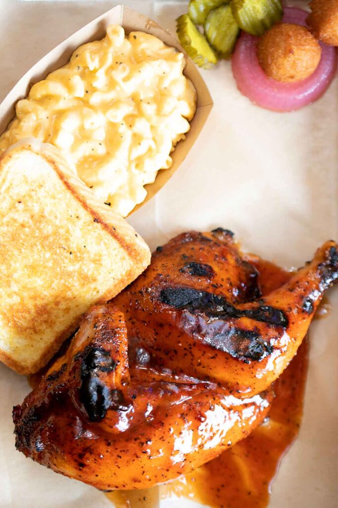 Tray of barbecued half chicken with toast and Mac n cheese, pickles on side.