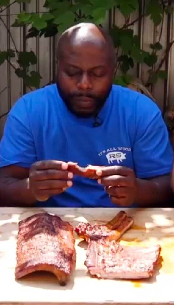 Rodney Scott sitting in front of a few racks of ribs, holding one showing the bite mark semicircle