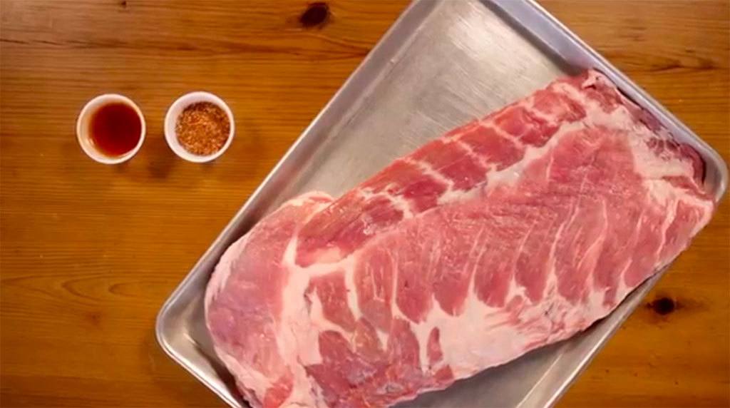 A rack of uncooked 10-12 bone spareribs on metal tray, with a cup of sauce and rub on the side