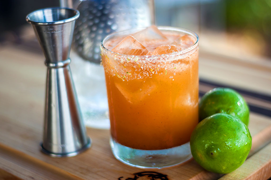 Mixed drink on wooden cutting board with limes and shot glass.