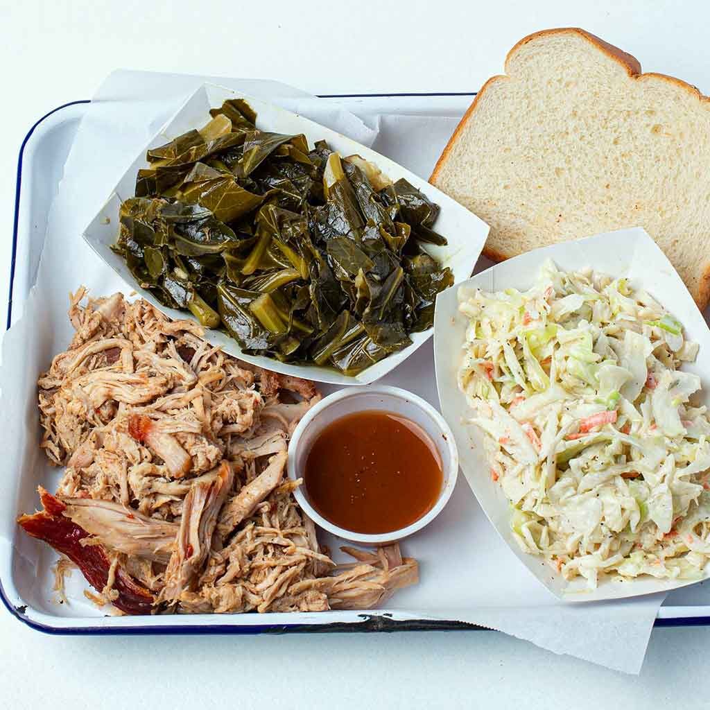 Pulled pork platter with collards, slaw, sauce, and white bread on metal tray with white butcher paper.
