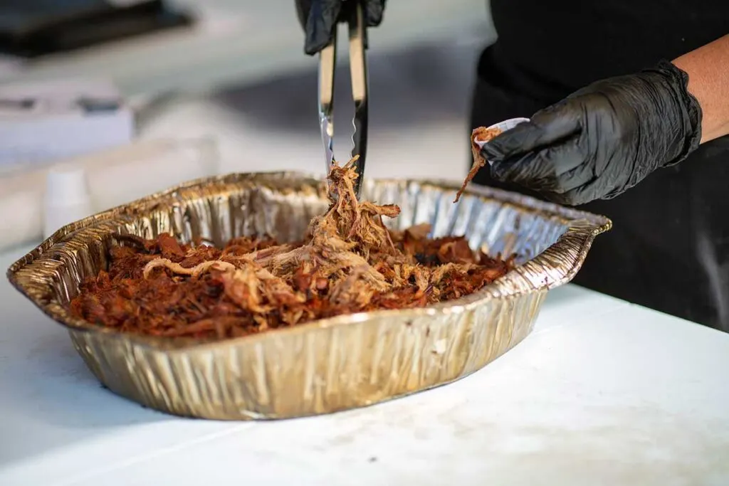 Tongs pull delicious looking pulled pork from dented, discolored foil pan