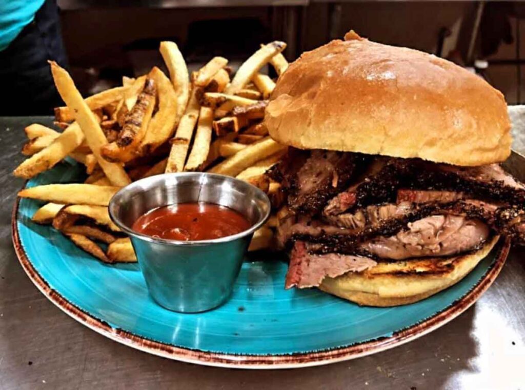 Brisket sandwich with fries on blue plate with a silver cup of ketchup