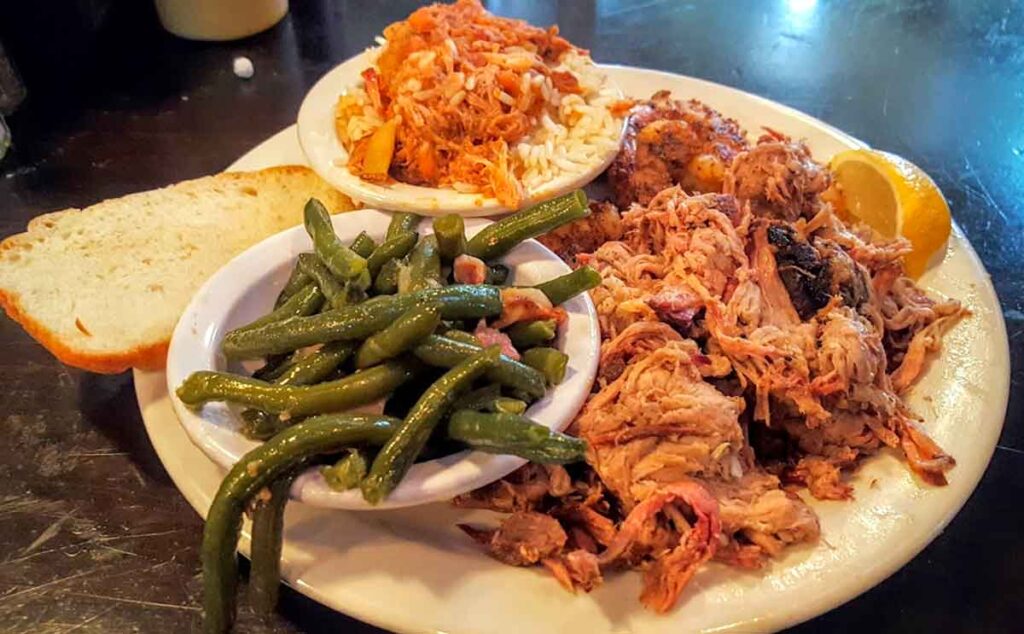 Plate on table at Q on Bay with small bowls of green beans and hash on rice, bread, pulled pork and a lemon.