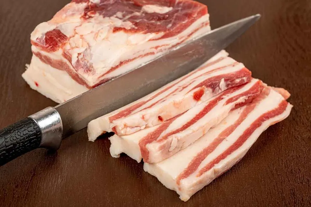 Knife cutting through large slabs of bacon from pork belly