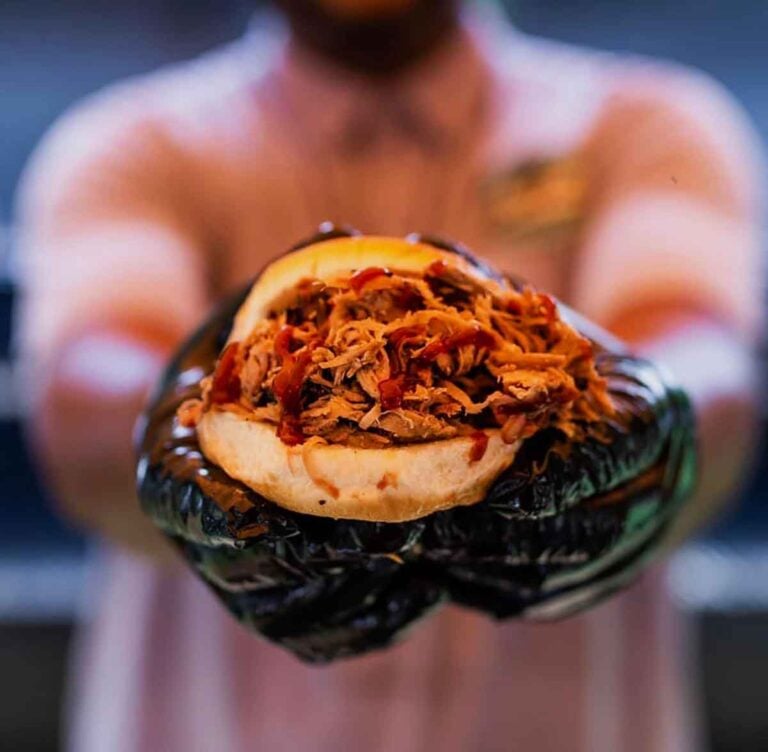 Gloved hands presenting a pulled pork sandwich on a regular bun.