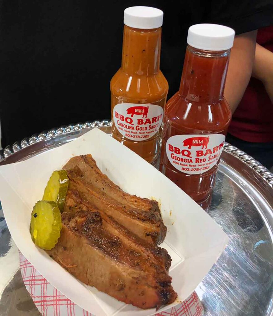 Paper tray with sliced brisket and pickles with a bottle of Carolina Gold sauce and Georgia Red sauce on silver platter.