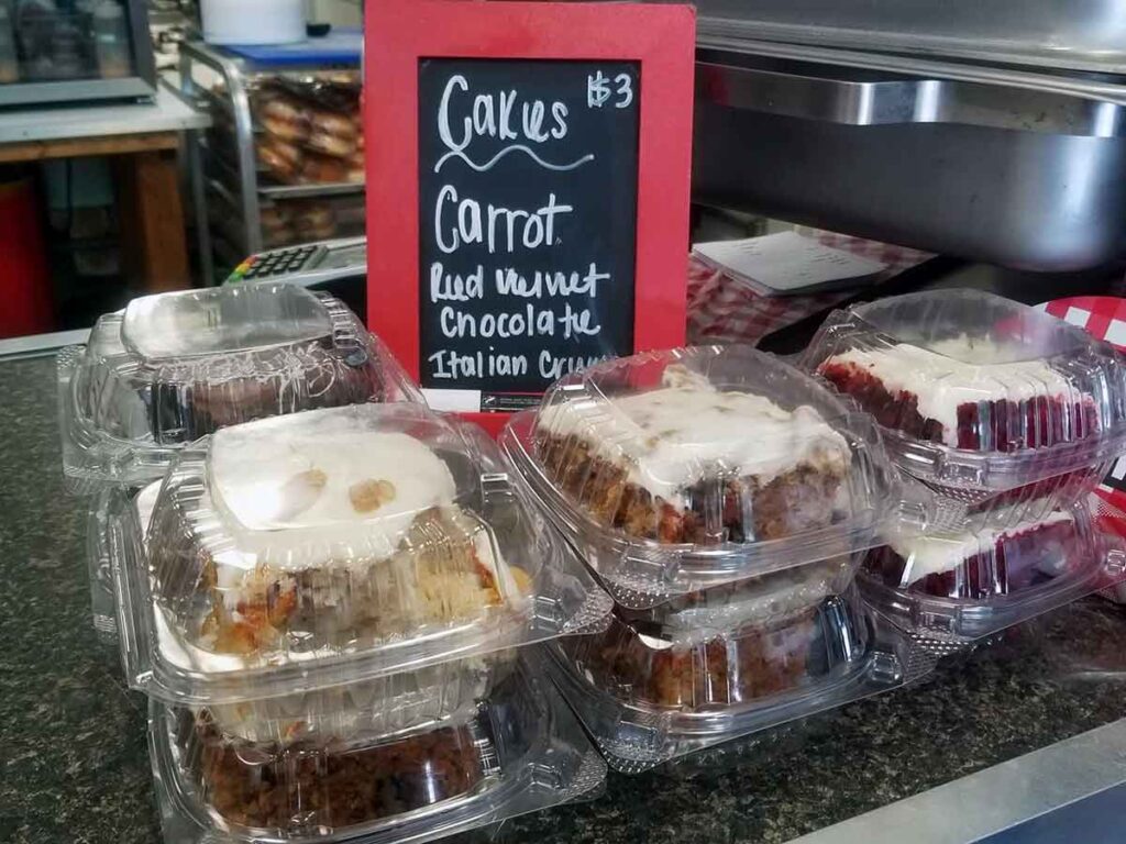 Plastic containers containing squares of cakes with a red framed chalkboard sign with types and price on a countertop.