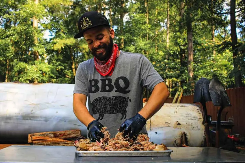 Hector Garate pulling pork on table outside with his pit behind him.