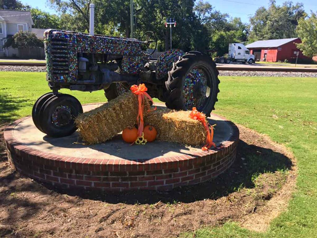 Tractor decorated and on round brick pedestal; bales of hay, pumpkins, and orange decorations in front.