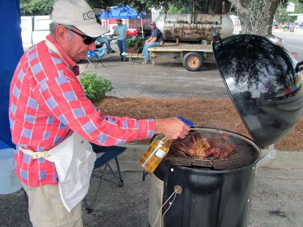 Older gentleman in a hat spraying meat on a grill with what looks like apple juice.