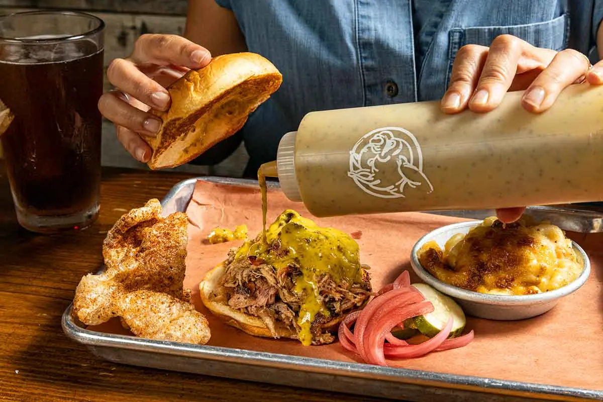 Closeup of hands pouring mustard sauce onto pulled pork sandwich