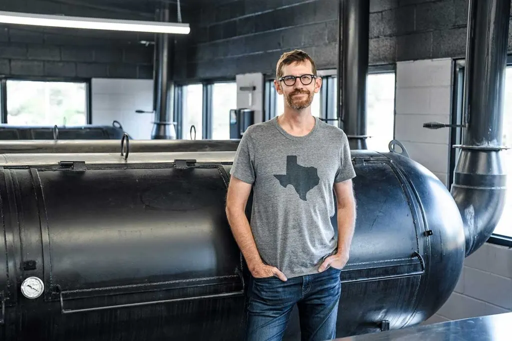 Wearing a gray t-shirt with a silhouette of Texas, John Lewis leans against one of his pits at Lewis Barbecue in Greenville