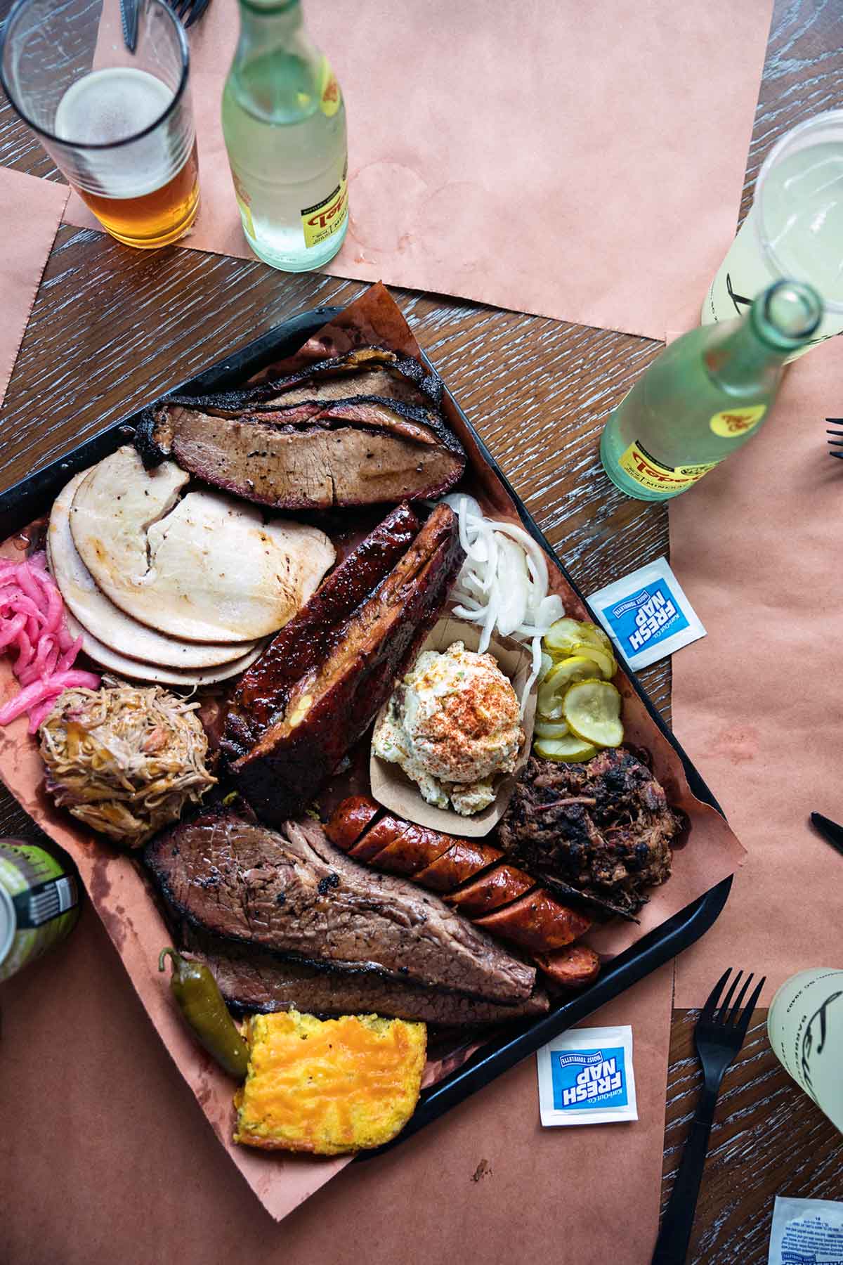 Overhead view of a tray on a wood table filled with meats and sides with drinks surrounding.