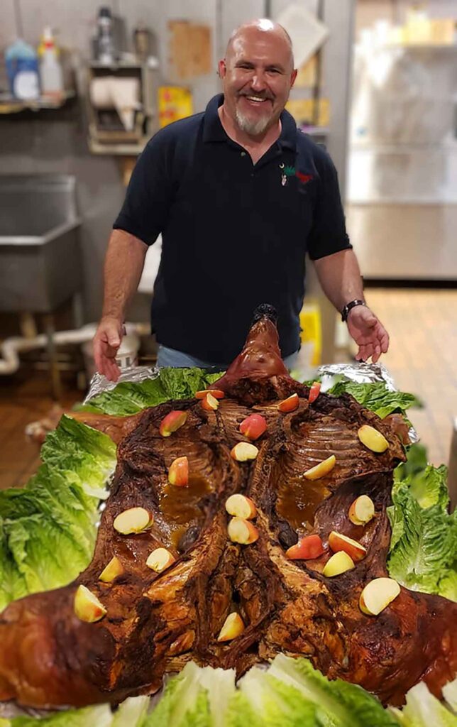 Bobby Griffin in front of smoked whole hog displayed on lettuce leaves and garnished with apple quarters.