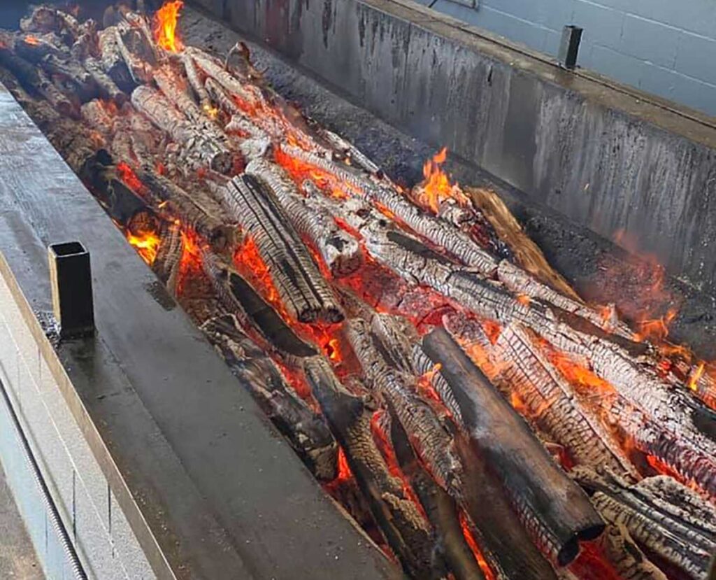 Pit filled with flaming logs burning down to embers to cook barbecue.
