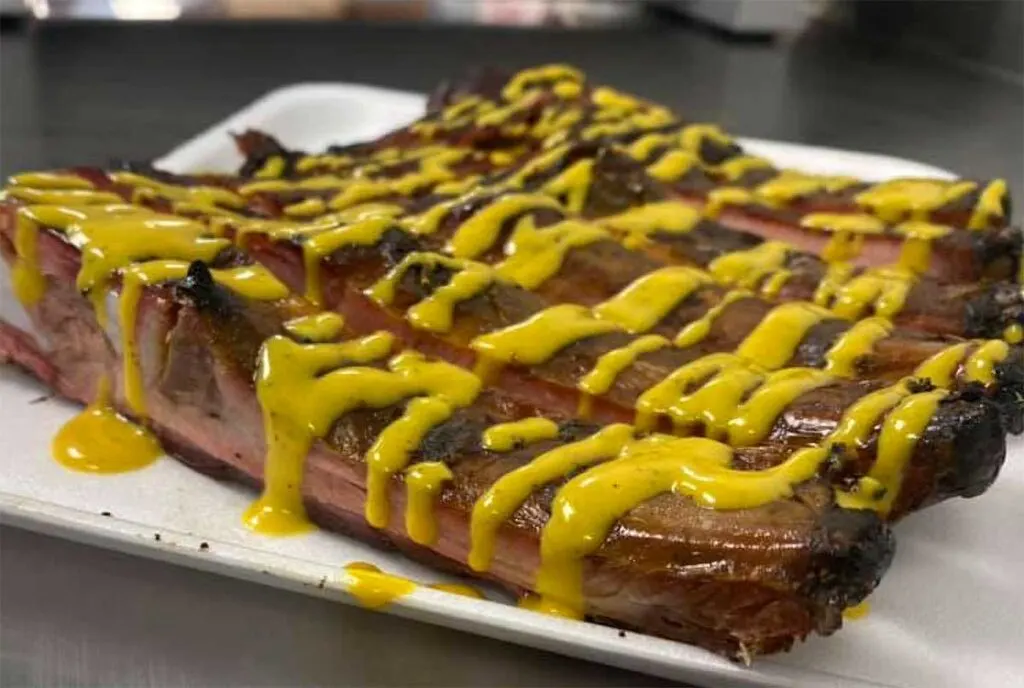 Plate of several smoked ribs drizzled with yellow mustard-based BBQ sauce.