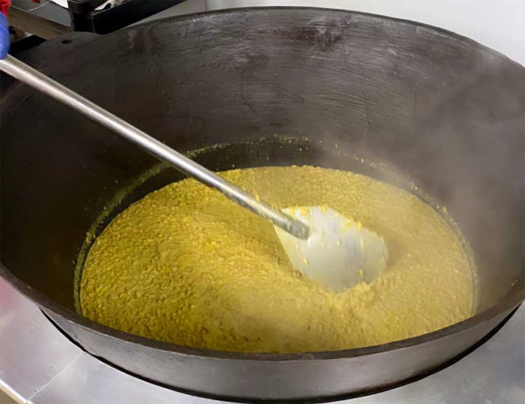 Yellow hash being stirred by hand in a large pot by a long metal oar-like instrument.