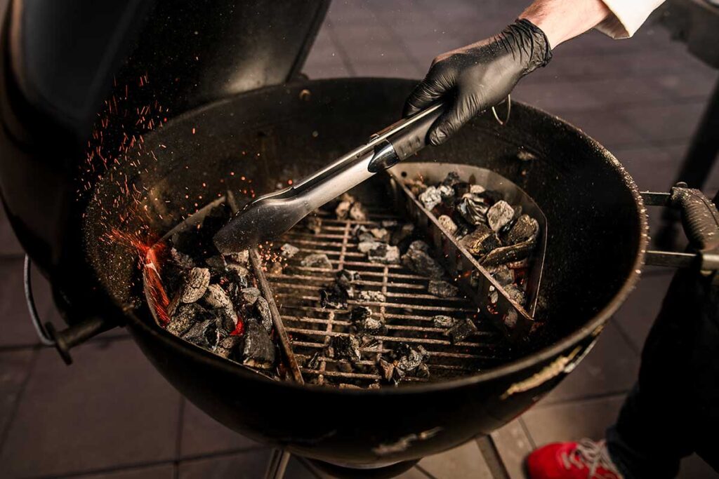 Hand with tongs moving hot charcoal to metal containers on the side of a Weber kettle grill.
