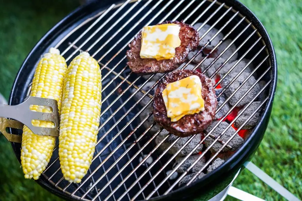 Two pieces of corn and two hamburgers with melting cheese on grill. Burgers are over a bank of hot coals.