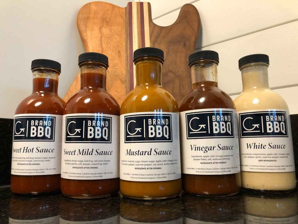 Bottles of sauce served by G Brand BBQ on black marble counter with a guitar shaped cutting board behind them.