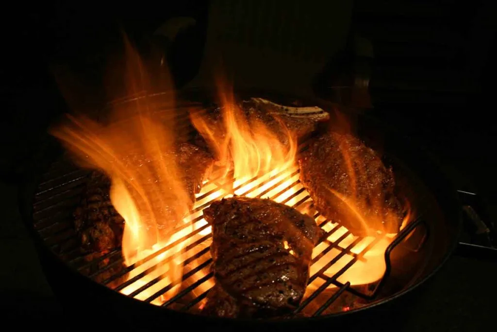 Flare-up from dripping fat around steaks on a charcoal kettle, showing direct heat over the coals and why two-zone grilling needs a cool side.