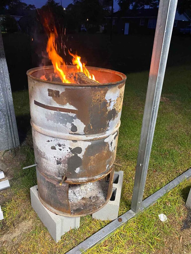 Burn barrel on concrete blocks with flames coming from the open top and embers in the shovel opening at the bottom.