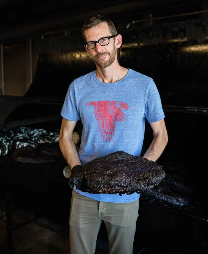 John Lewis in front of pit holding finished brisket with great bark.