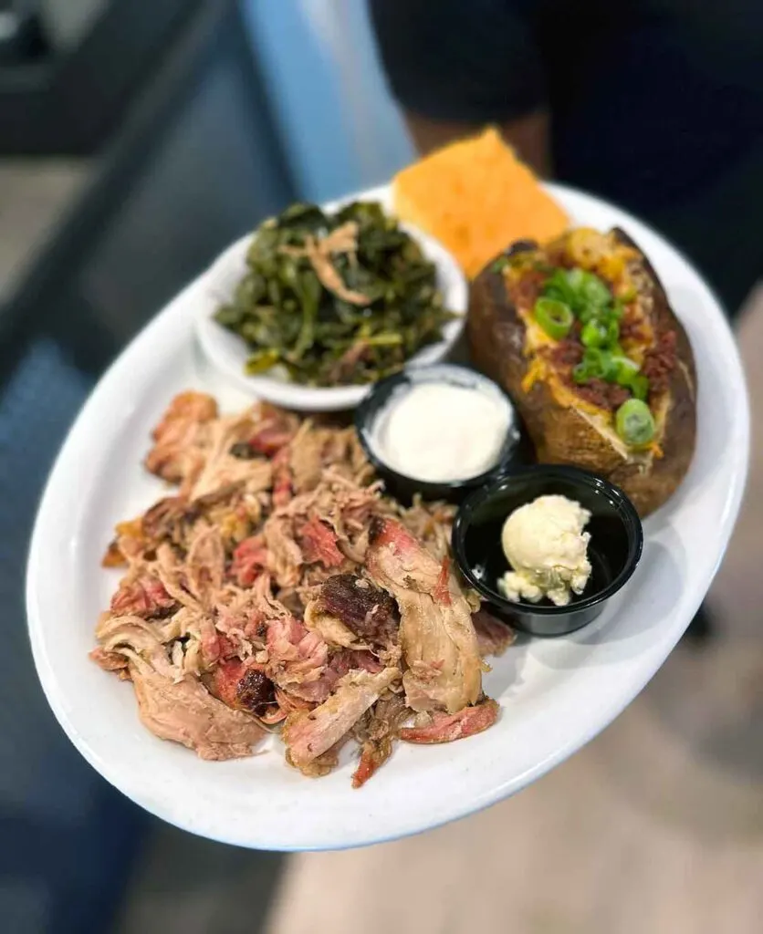 Pulled pork, collards, cornbread, and a loaded baked potato on plate with containers of butter and sour cream.