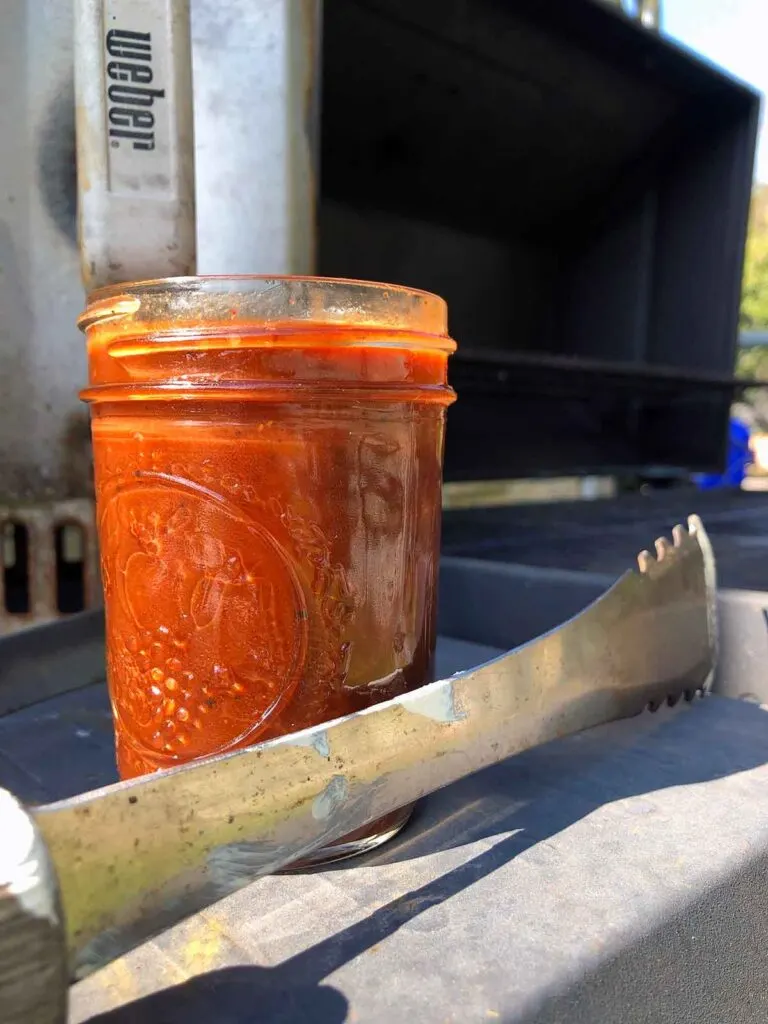 Tongs, mason jar of red BBQ sauce, and Weber Rapidfire chimney starter on grill shelf.