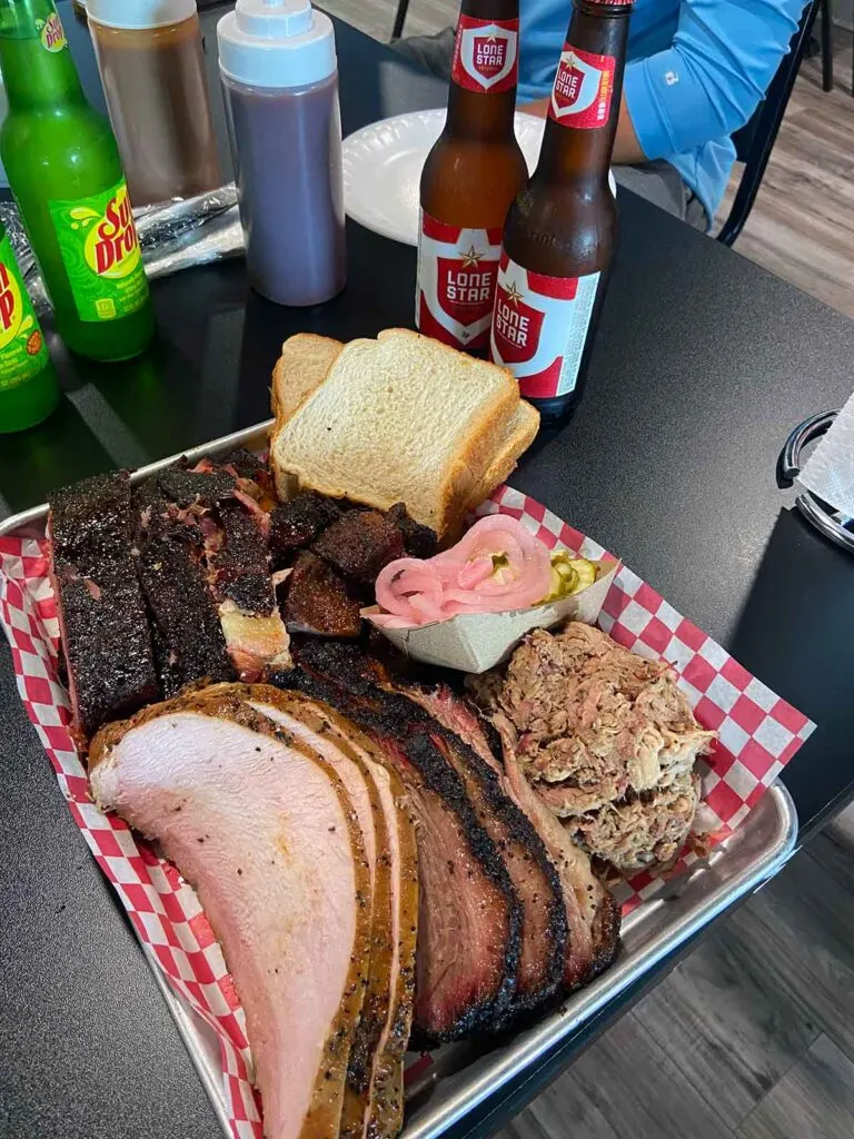 Tray of meats and sides on table with sauces and drinks.