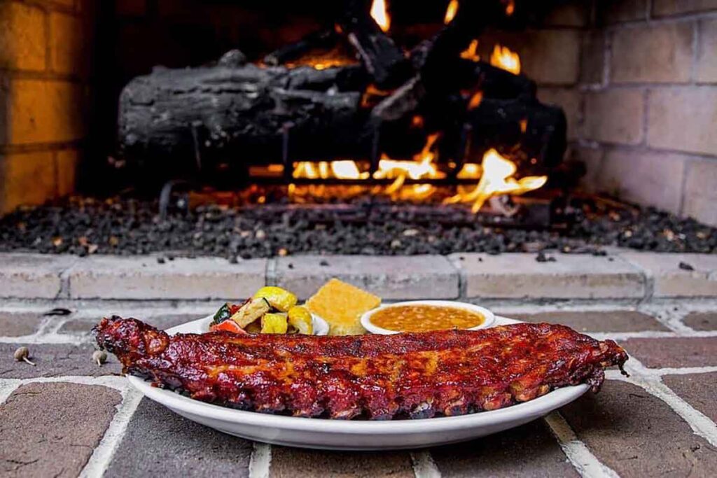 Plate with a slab of ribs, fire roasted vegetables, cornbread, and baked beans on hearth in front of a fire.