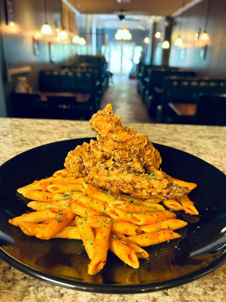 Wings over pasta on black plate with restaurant interior in background.