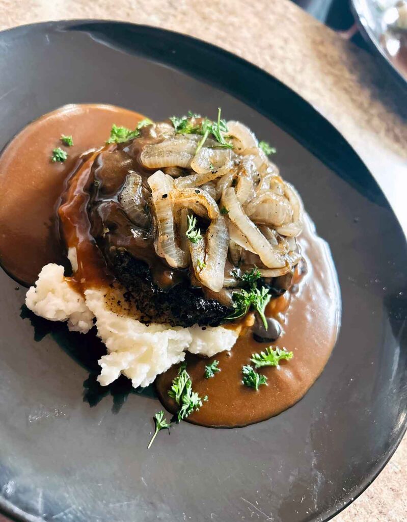Black plate with base of brown gravy and mashed potatoes toped with hamburger steak and sautéed onions.