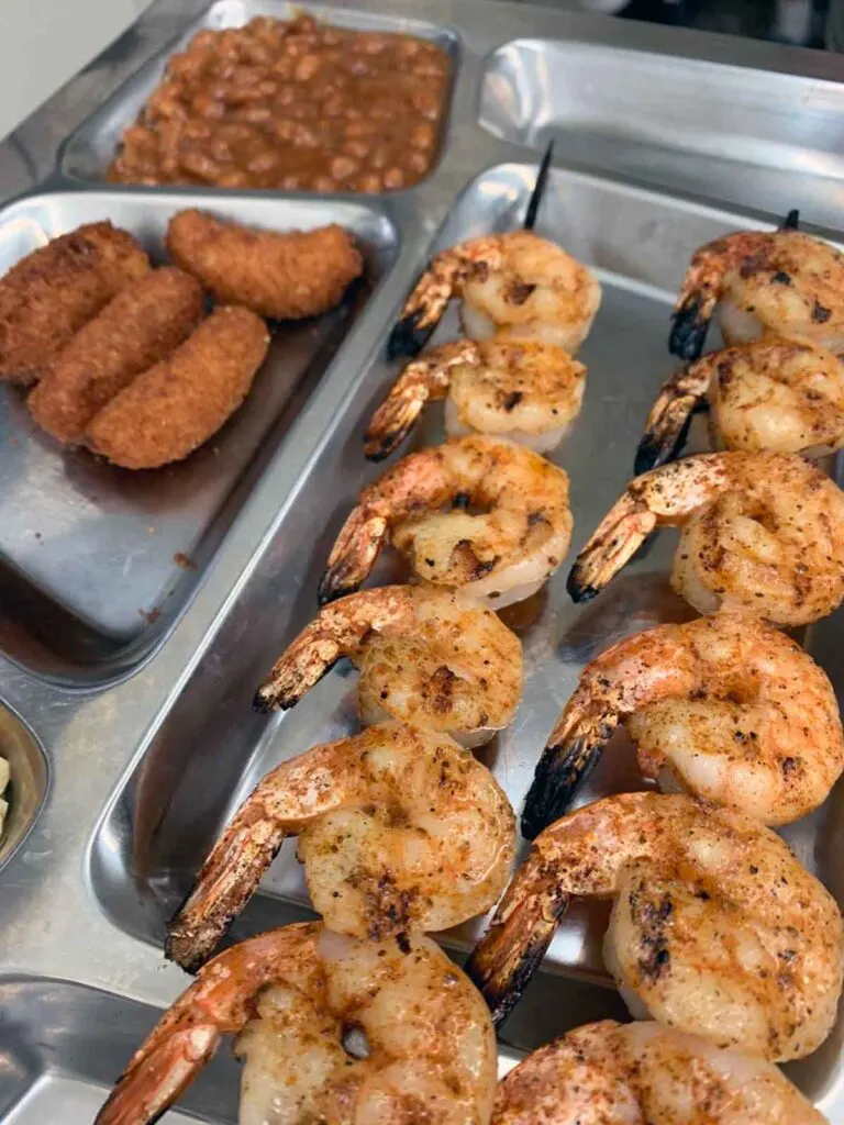 Two skewers of grilled shrimp on metal tray with hush puppies and baked beans.