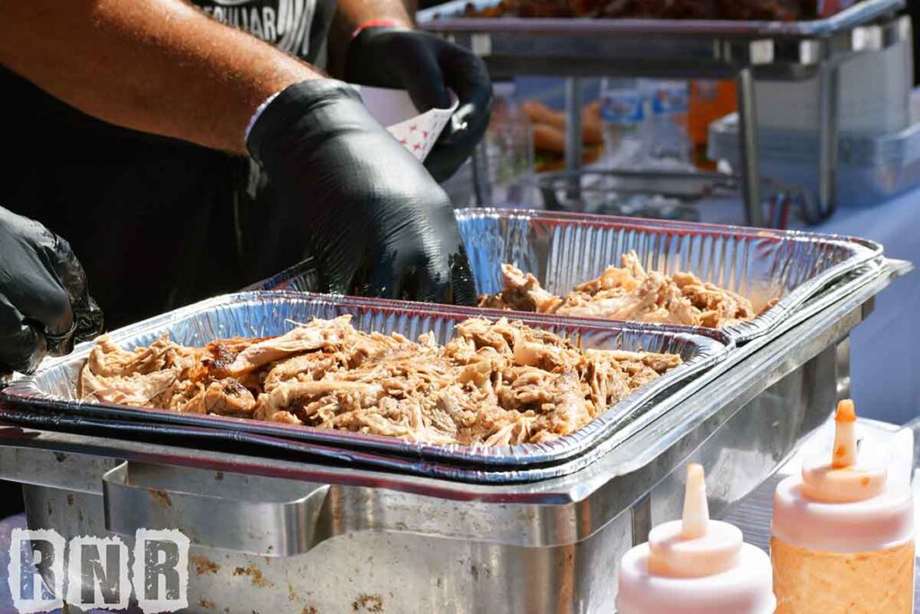 A gloved hand reaching into pans of pulled pork to serve.