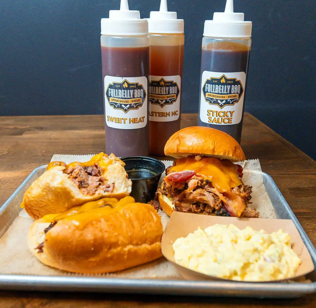 Tray of BBQ sandwiches, potato salad, and dipping sauce in front of three bottles of Fullbelly BBQ's sauces.