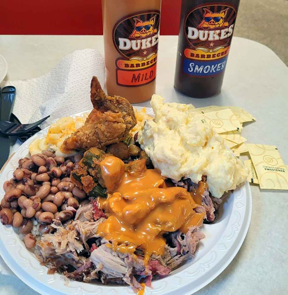 Plate with BBQ sauce covered BBQ, chicken and sides with sauce bottles in background.